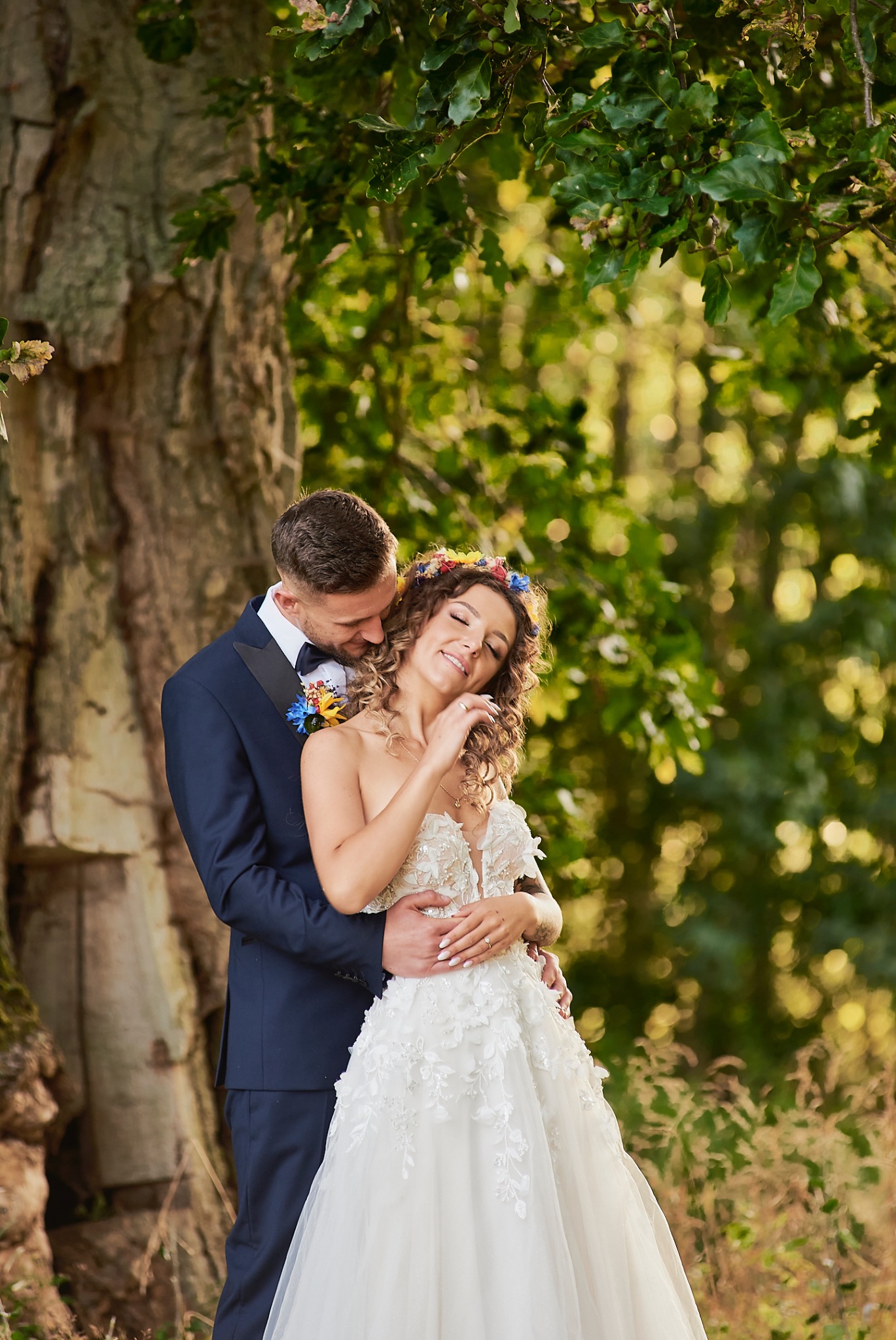 Uśmiechnięta panna młoda w koronie z kwiatów i zwiewnej sukni ślubnej, obejmowana przez pana młodego w granatowym smokingu na tle drzewa.