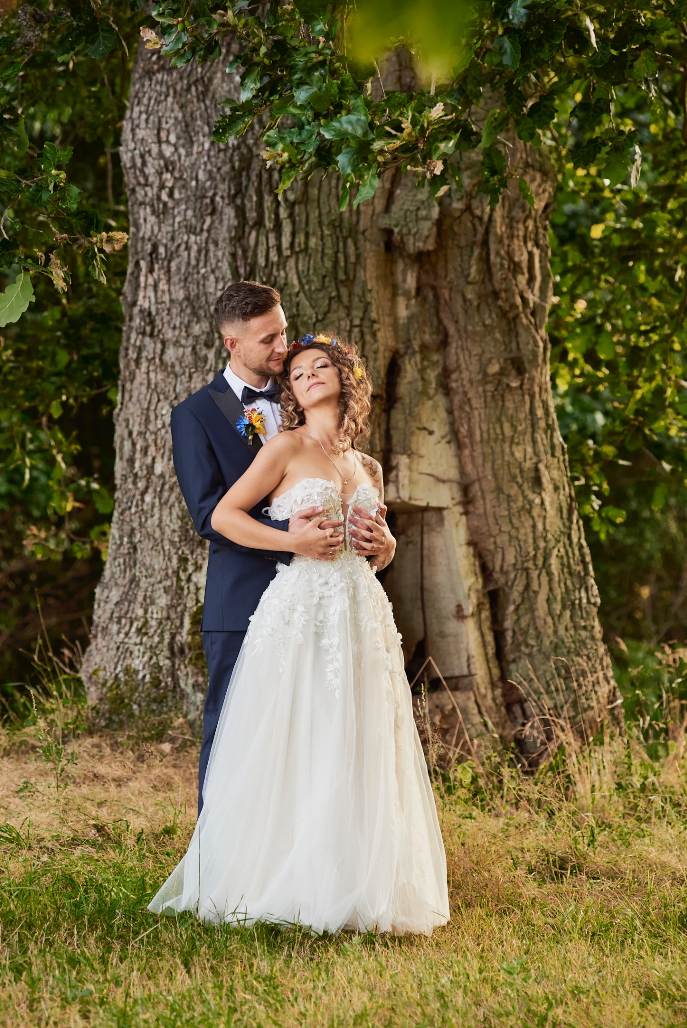 Para młoda: Panna młoda w koronkowej sukni bez ramiączek i wianku z kwiatów, obejmowana przez pana młodego w granatowym garniturze z muchą, pozuje pod rozłożystym drzewem w naturalnym świetle.