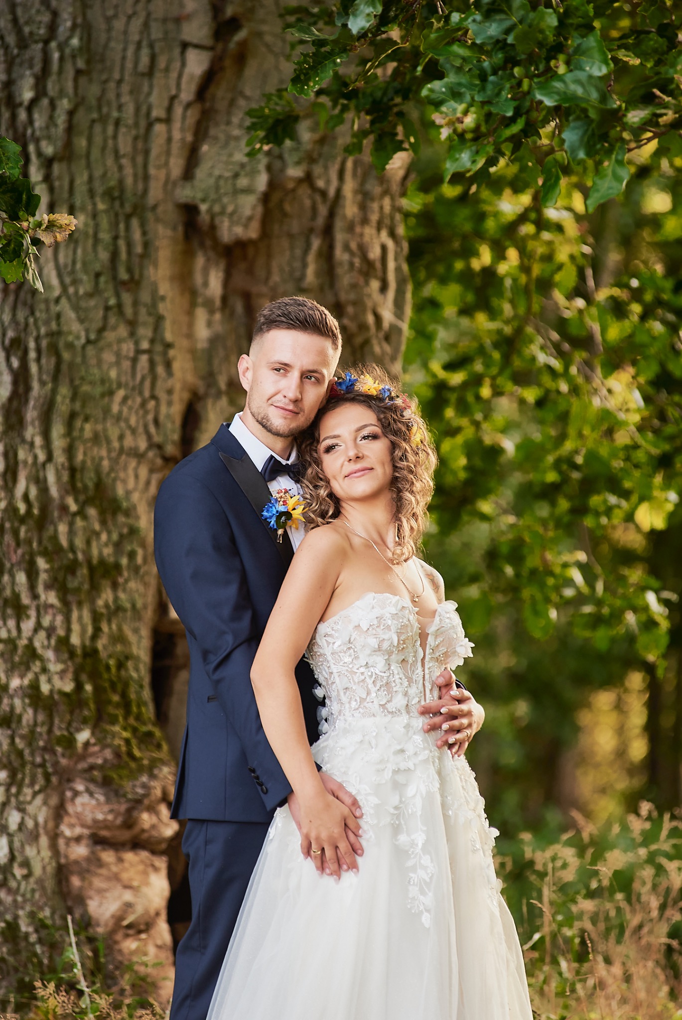 Para młoda w lesie: panna młoda w białej sukni z kwiatowymi aplikacjami i wiankiem na głowie, pan młody w granatowym smokingu z muchą, pozują przy drzewie.