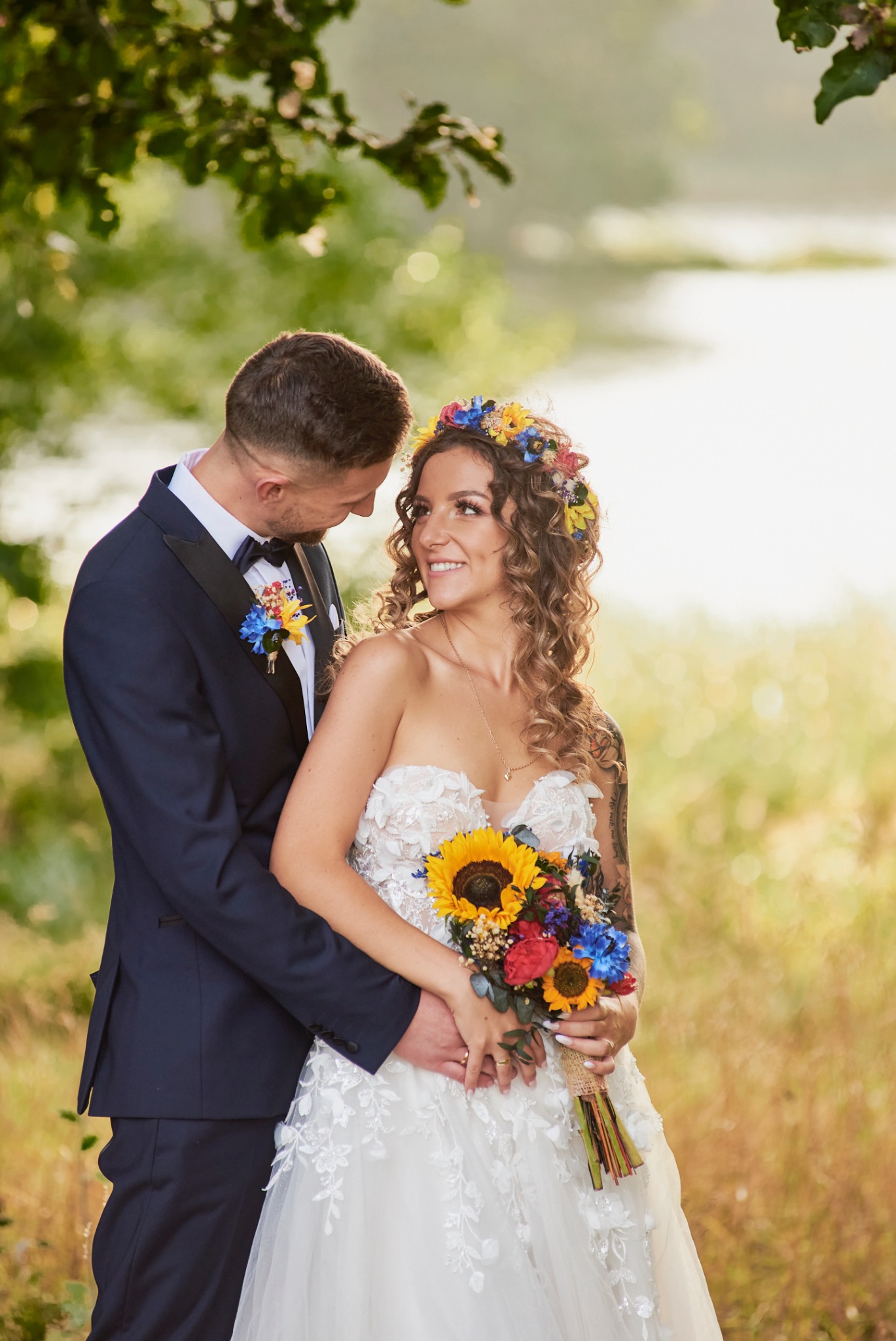 Uśmiechnięta panna młoda w sukni z koronkowymi aplikacjami i wiankiem z polnych kwiatów trzyma bukiet słoneczników, obejmowana przez pana młodego w granatowym smokingu z muchą i butonierką.