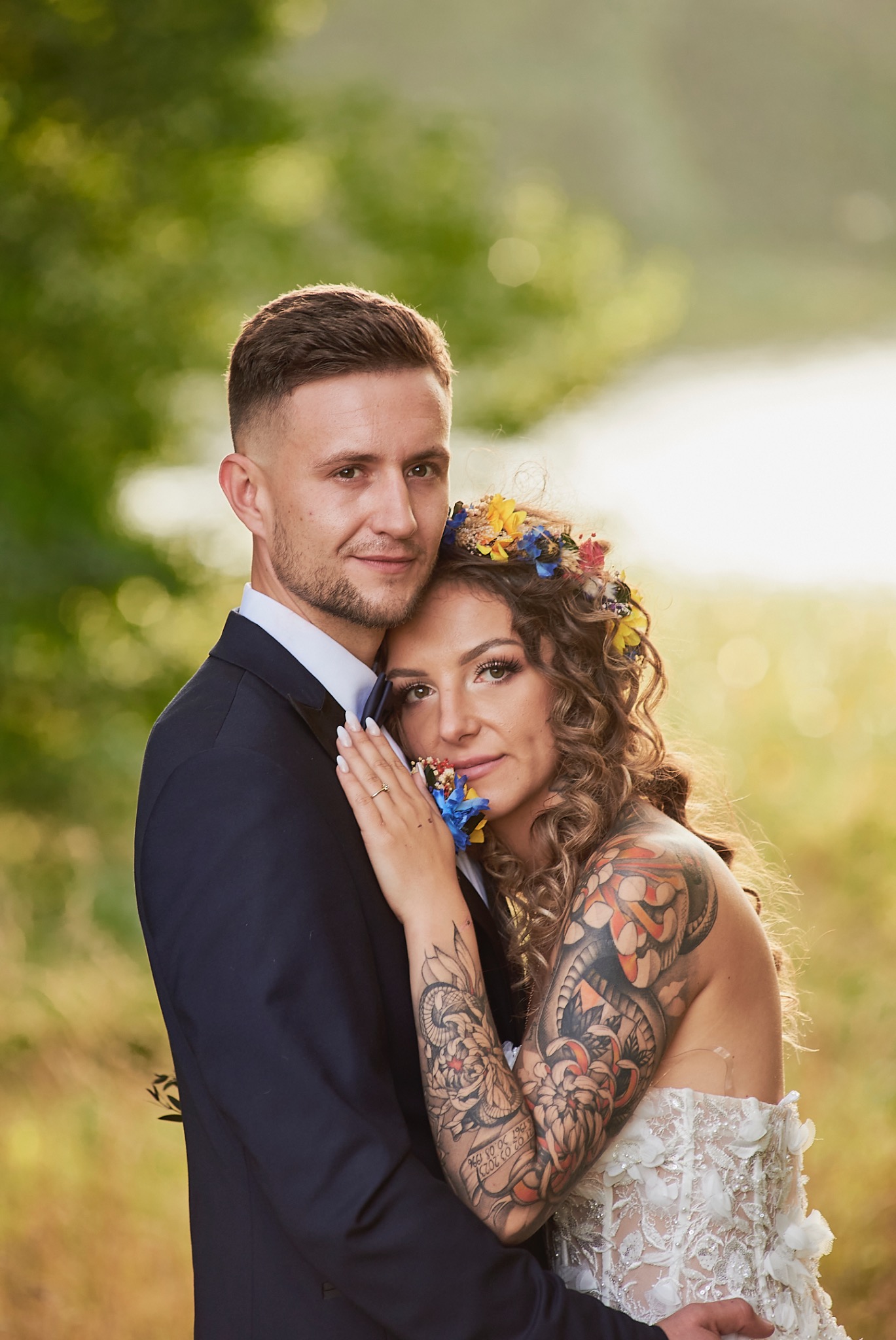 Uśmiechnięta para młoda w objęciach na tle zieleni; Panna młoda z tatuażem na ramieniu i wiankiem z polnych kwiatów.