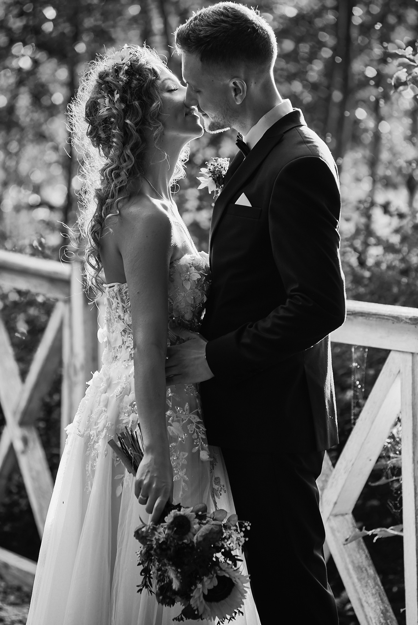 Czarno-białe zdjęcie całującej się pary młodej na tle drewnianej balustrady; panna młoda w sukni z kwiatowymi aplikacjami trzyma bukiet słoneczników.