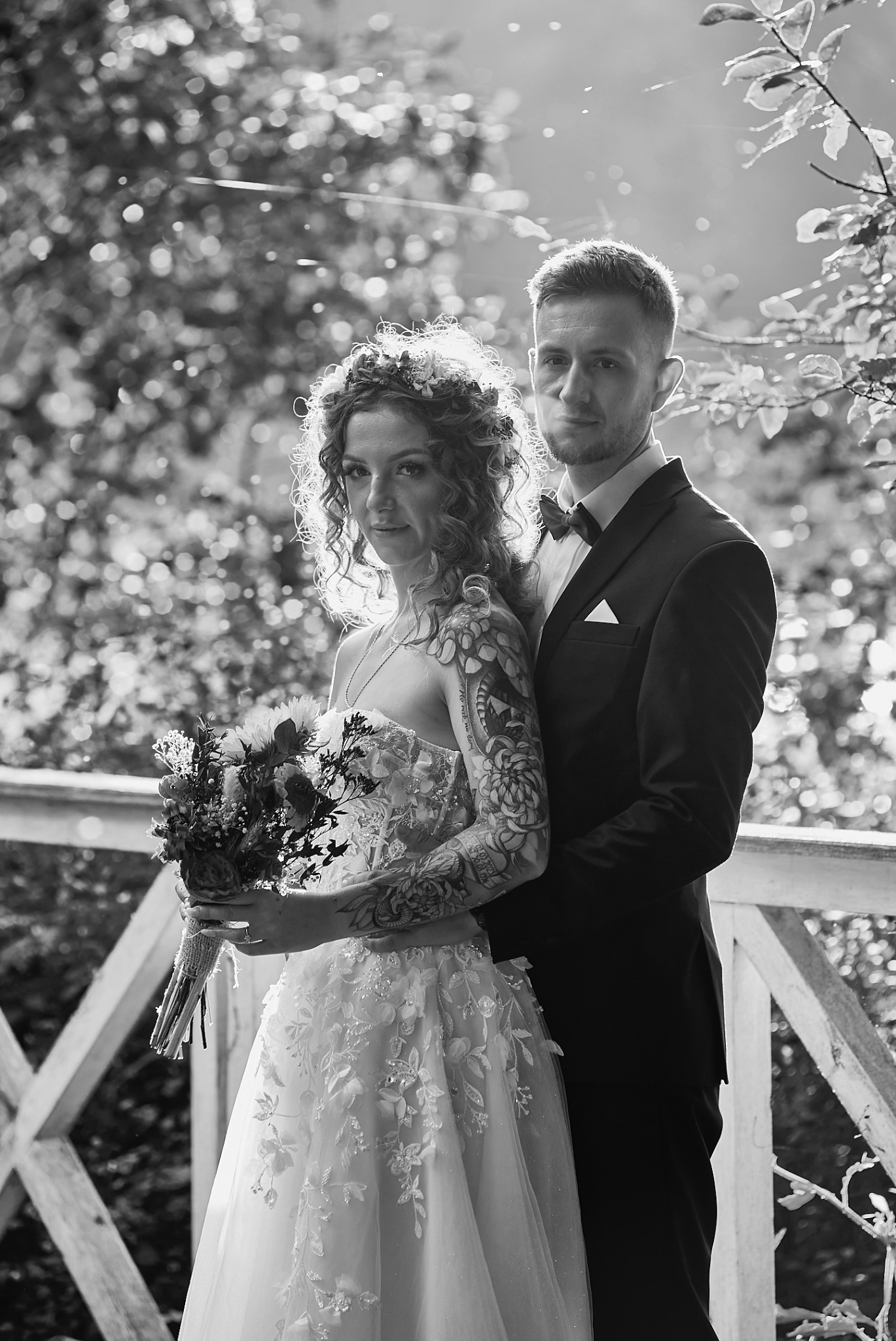 Czarno-białe zdjęcie pary młodej: panna młoda z tatuażami na ramieniu trzyma bukiet, obok pan młody w garniturze pod drewnianą balustradą, z promieniami słońca w tle.