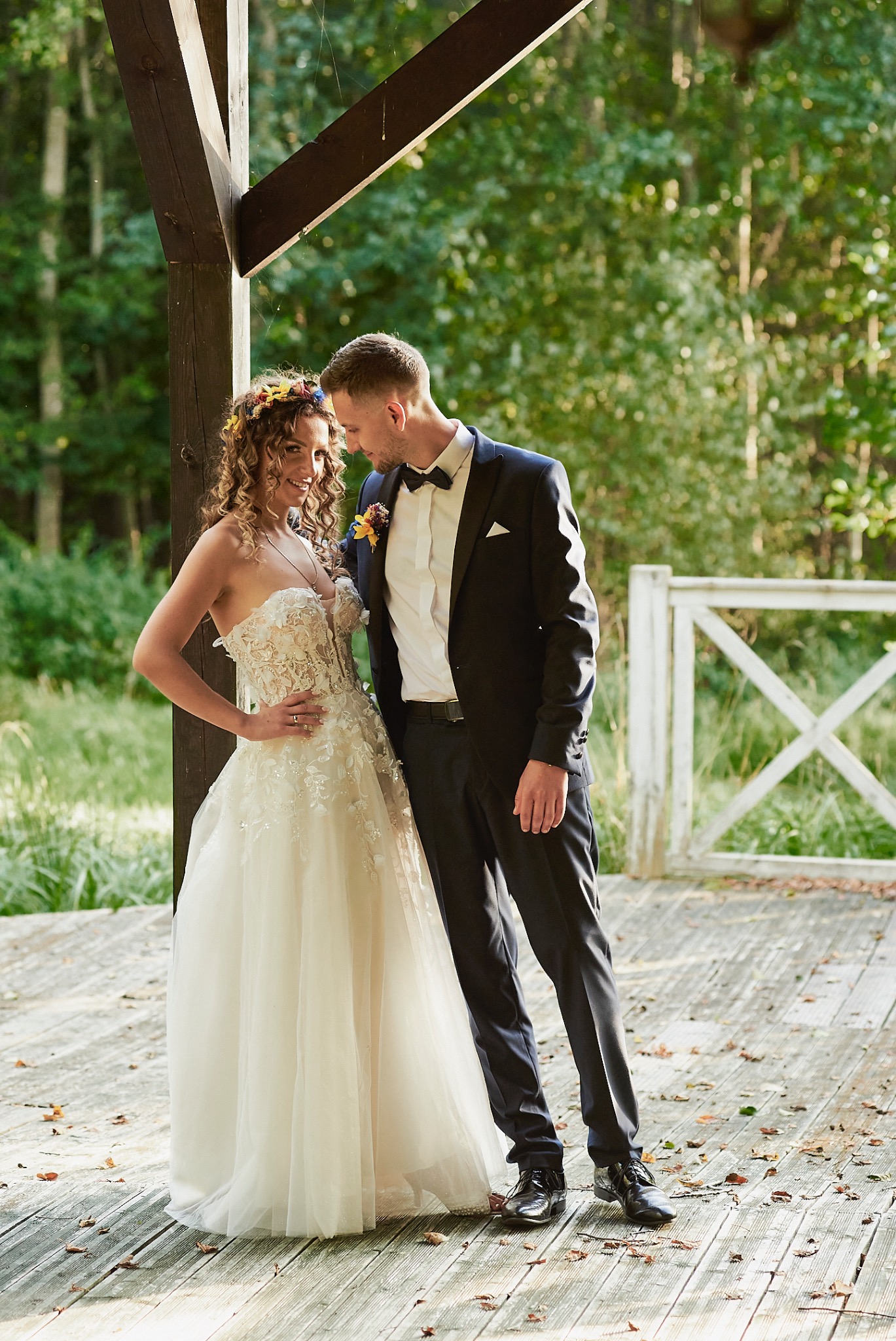 Para młoda w plenerze: panna młoda w sukni bez ramiączek z kwiatowym haftem i wiankiem, pan młody w czarnym garniturze i muszce, stoją na drewnianym podeście z opadłymi liśćmi, w tle zielony las...