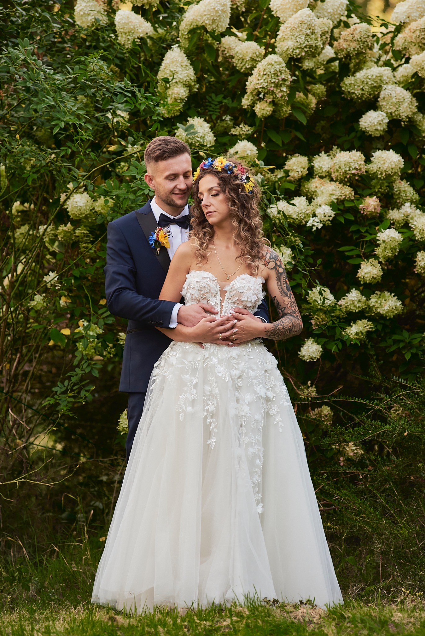 Para młoda: panna młoda w sukni z aplikacjami kwiatowymi i wiankiem, pan młody w granatowym smokingu, obejmująca się na tle kwitnących hortensji.