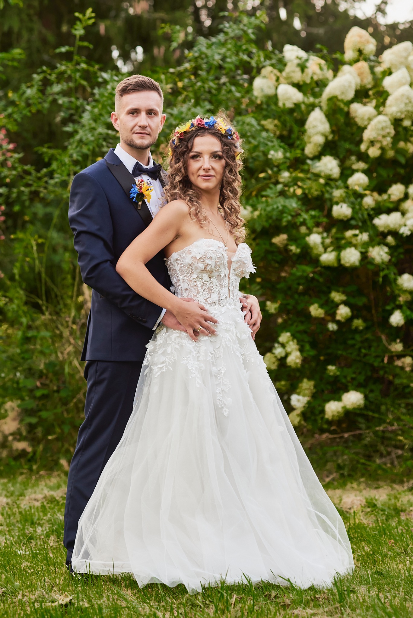 Para młoda w plenerze: Panna młoda w koronkowej sukni z kwiatowym wiankiem, pan młody w granatowym smokingu z butonierką, pozują na tle kwitnących krzewów.