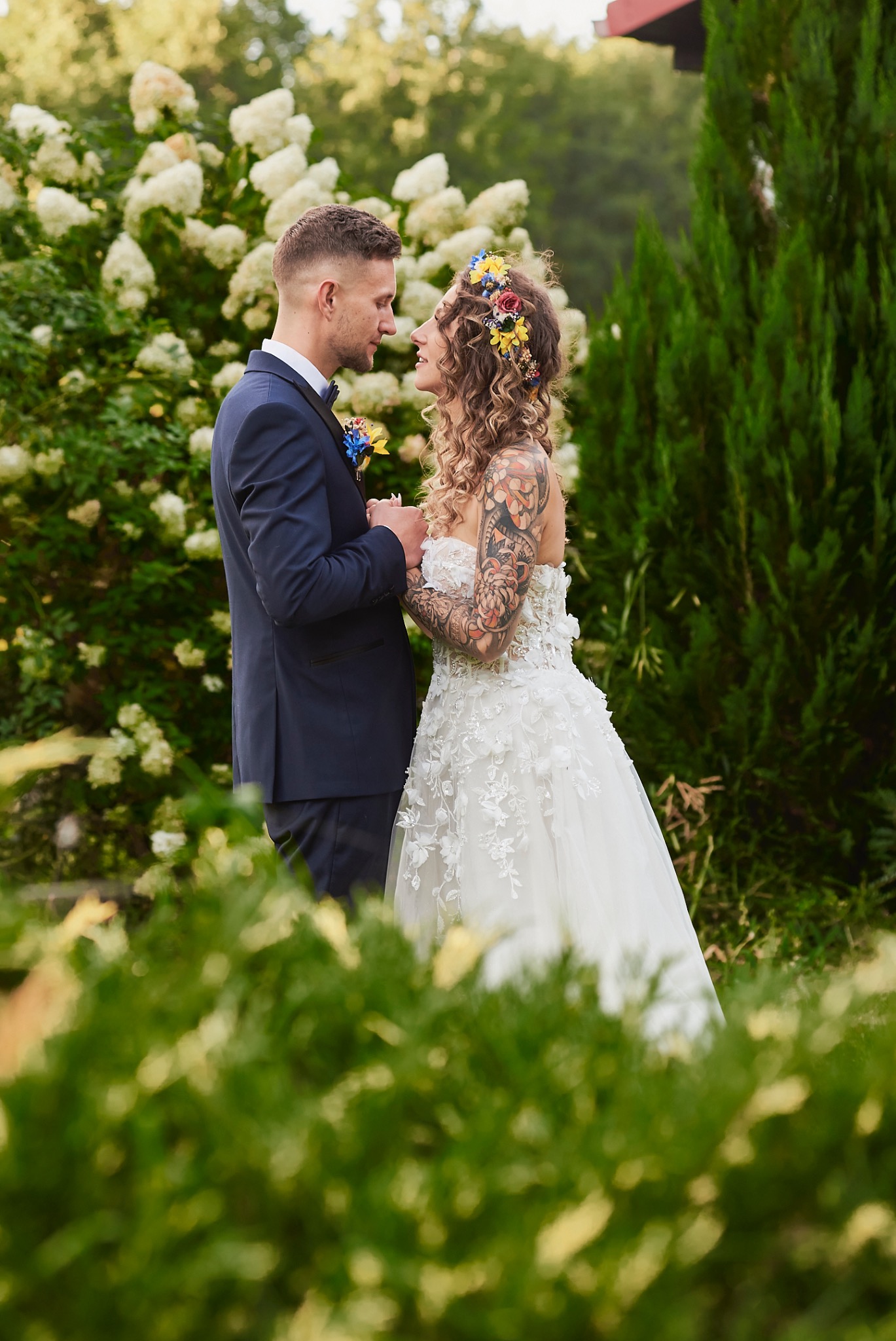 Para młoda w ogrodzie: panna młoda w białej sukni z kwiatowymi aplikacjami i wiankiem z kolorowych kwiatów, pan młody w granatowym garniturze z butonierką, trzymają się za ręce, patrząc na siebie.