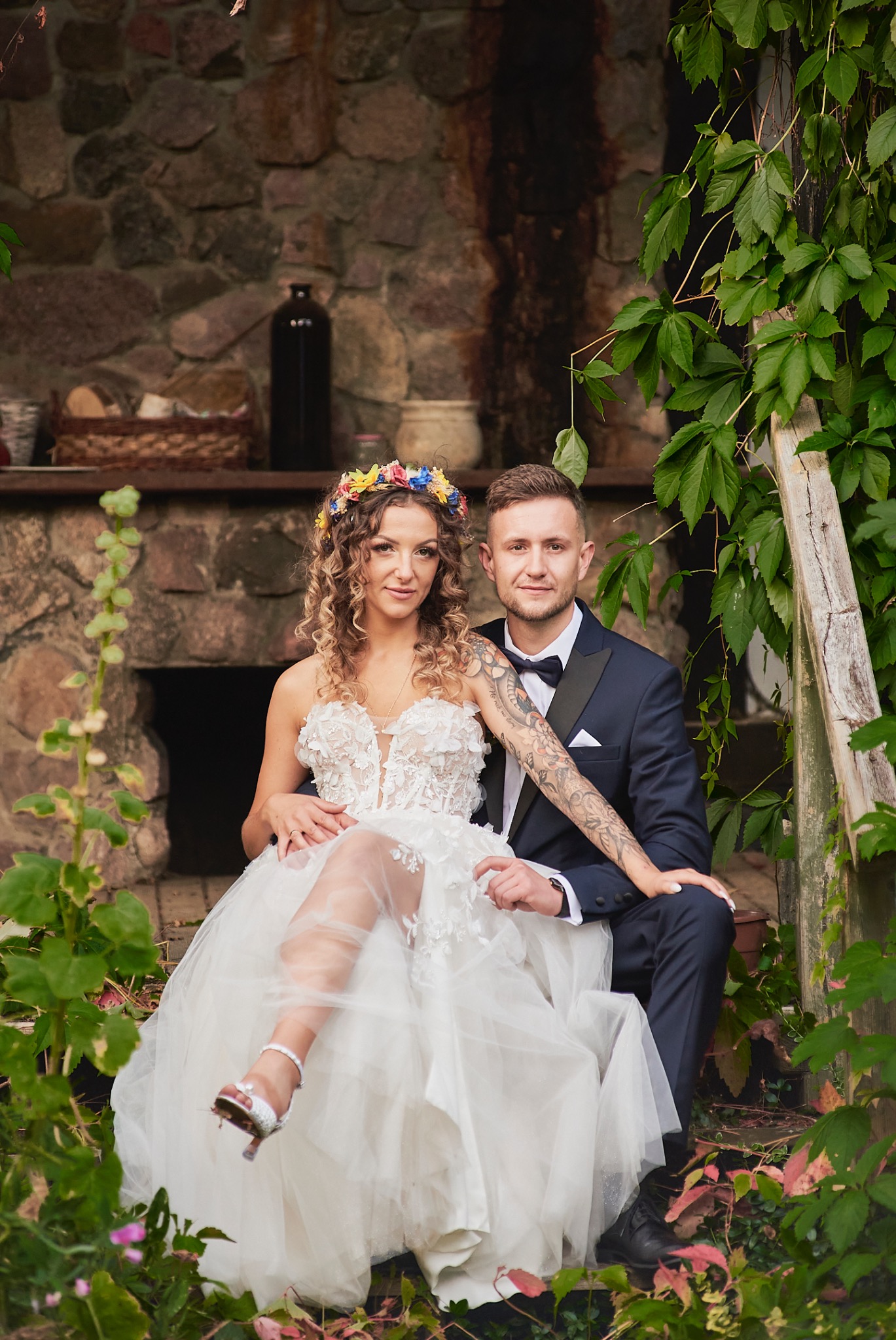 Para młoda siedzi na schodach przed kamiennym budynkiem, panna młoda w białej sukni z kwiatowym wiankiem, pan młody w granatowym garniturze z muszką i tatuażem na ramieniu.