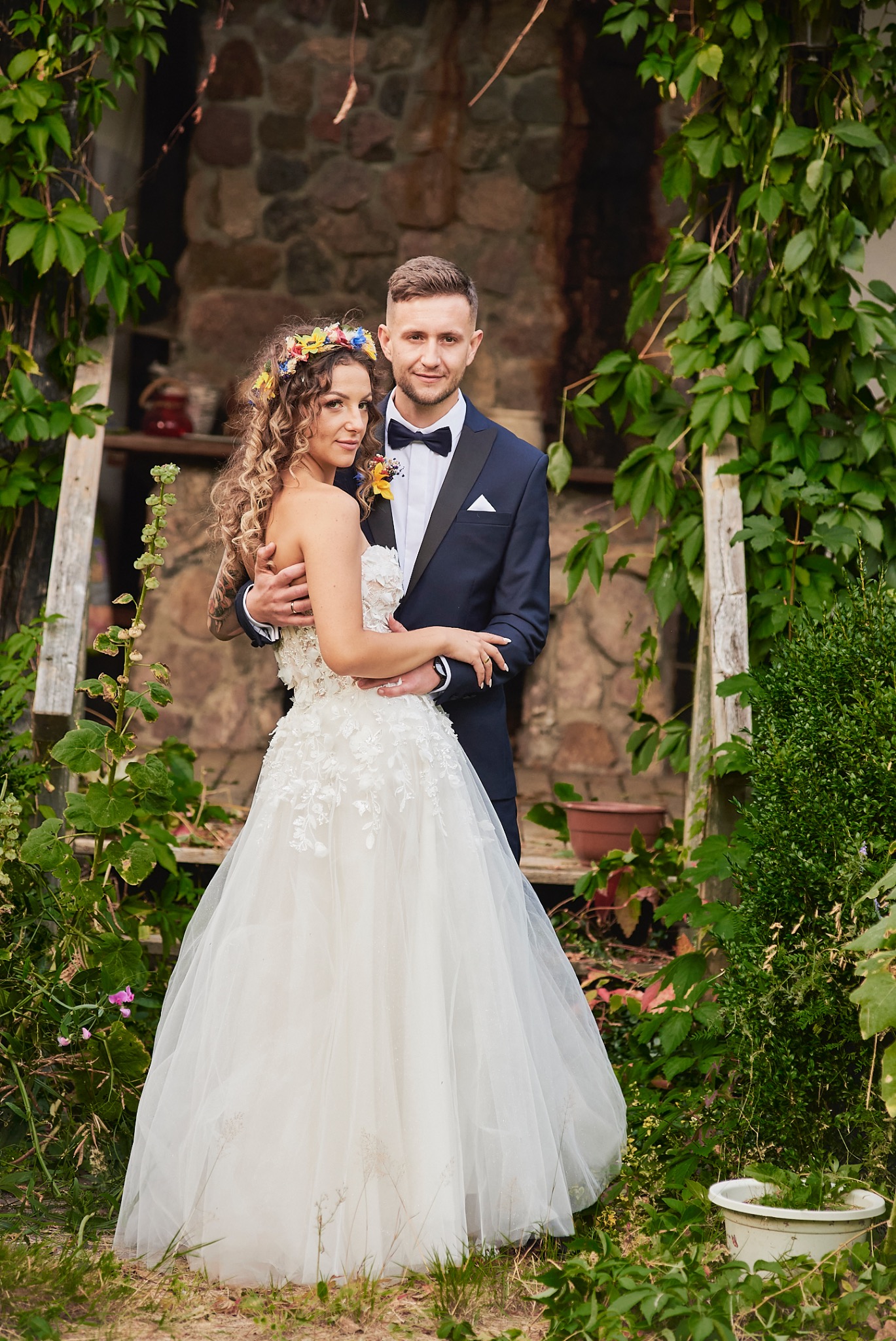 Para młoda: panna młoda w białej sukni z kwiatowymi aplikacjami i wiankiem, pan młody w granatowym smokingu, pozują na tle kamiennej ściany porośniętej bluszczem i drewnianych schodów.