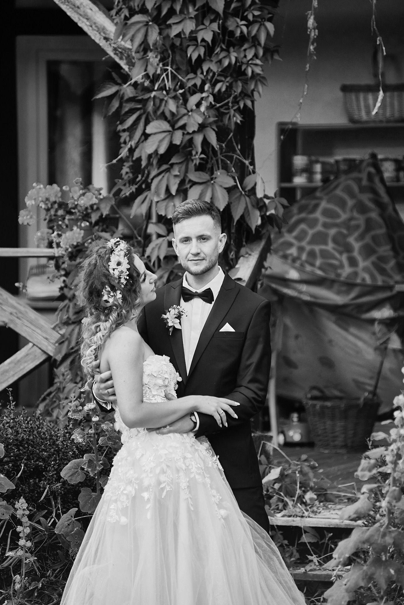 Czarno-białe zdjęcie pary młodej w objęciach; panna młoda w sukni z kwiatowym wzorem i wiankiem na głowie, pan młody w smokingu z muszką, oboje otoczeni zielenią.