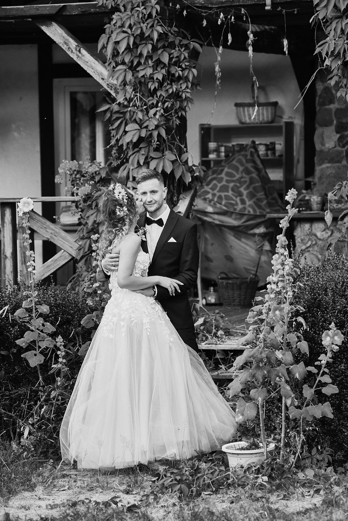 Czarno-białe zdjęcie pary młodej w Miastku: panna młoda w koronkowej sukni i wianku, pan młody w smokingu z muszką, obejmujący się na tle porośniętej zielenią werandy.