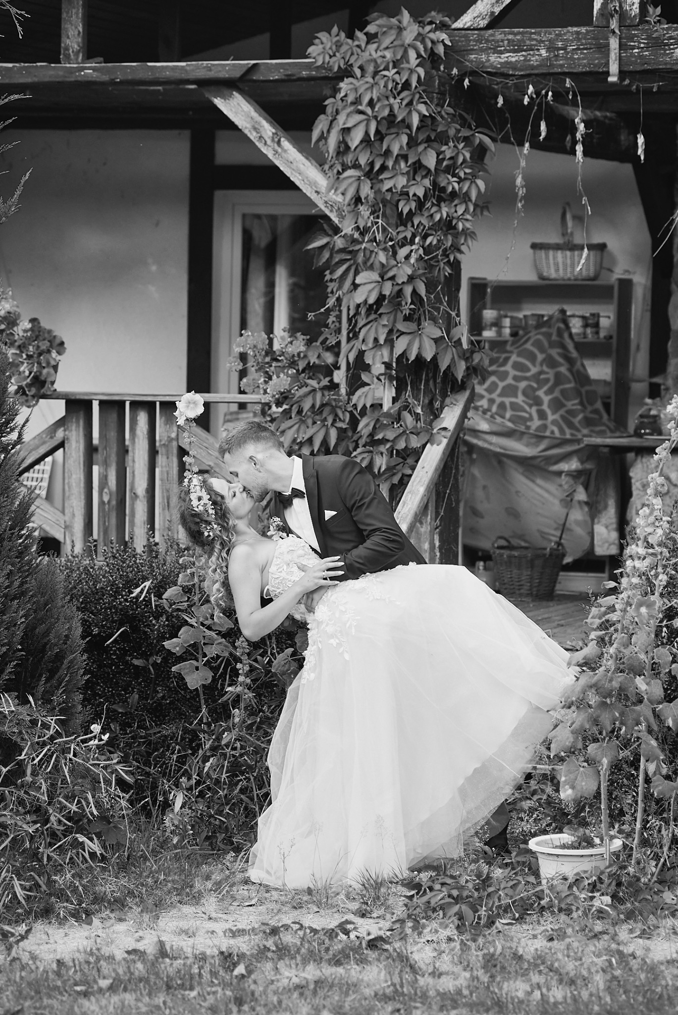 Czarno-białe zdjęcie całującej się pary młodej na tle drewnianego domu porośniętego bluszczem, panna młoda w koronkowej sukni i wianku, pan młody w smokingu.