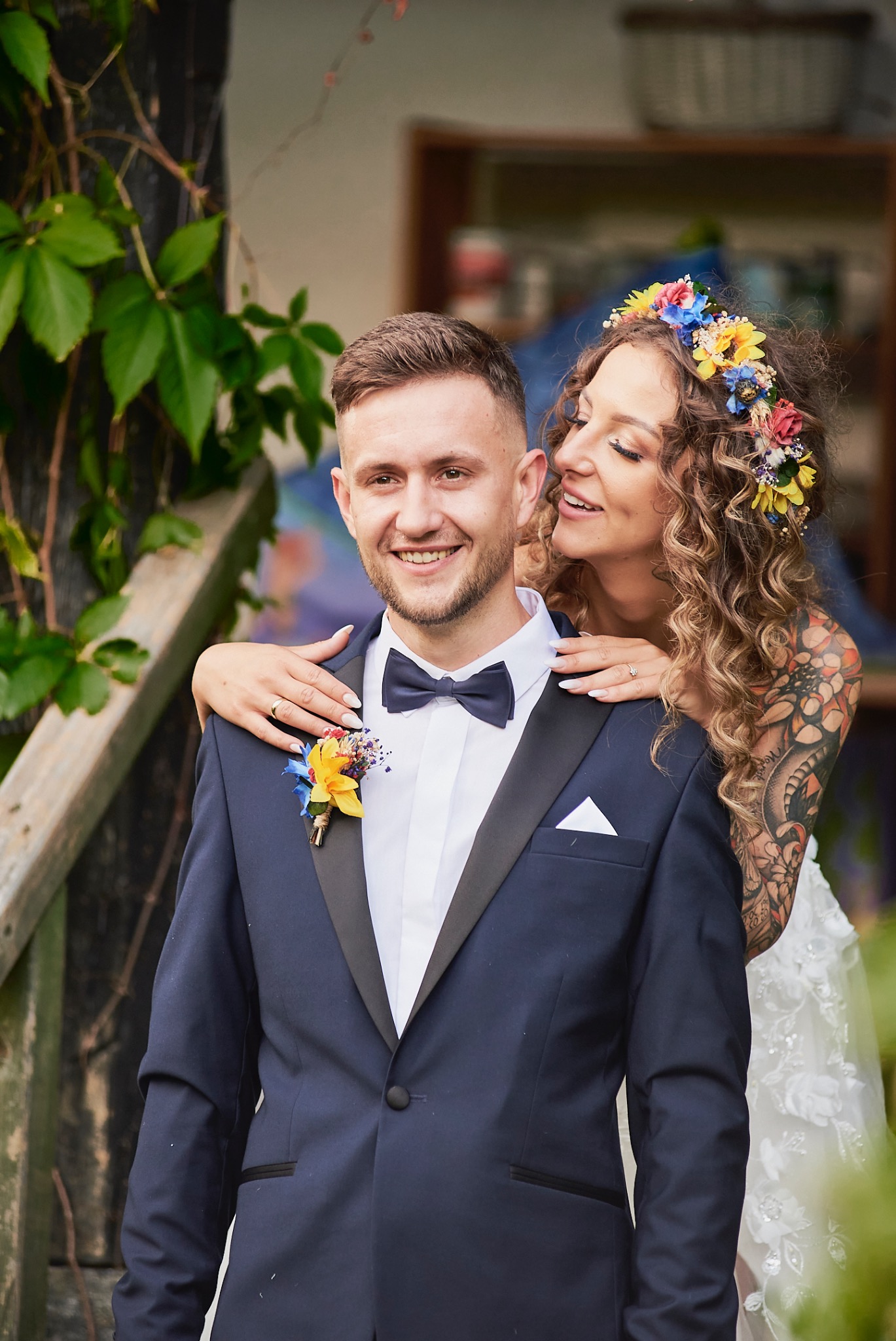 Uśmiechnięty pan młody w granatowym smokingu z butonierką, obejmowany przez pannę młodą z kwiatowym wiankiem i tatuażami na ramieniu.