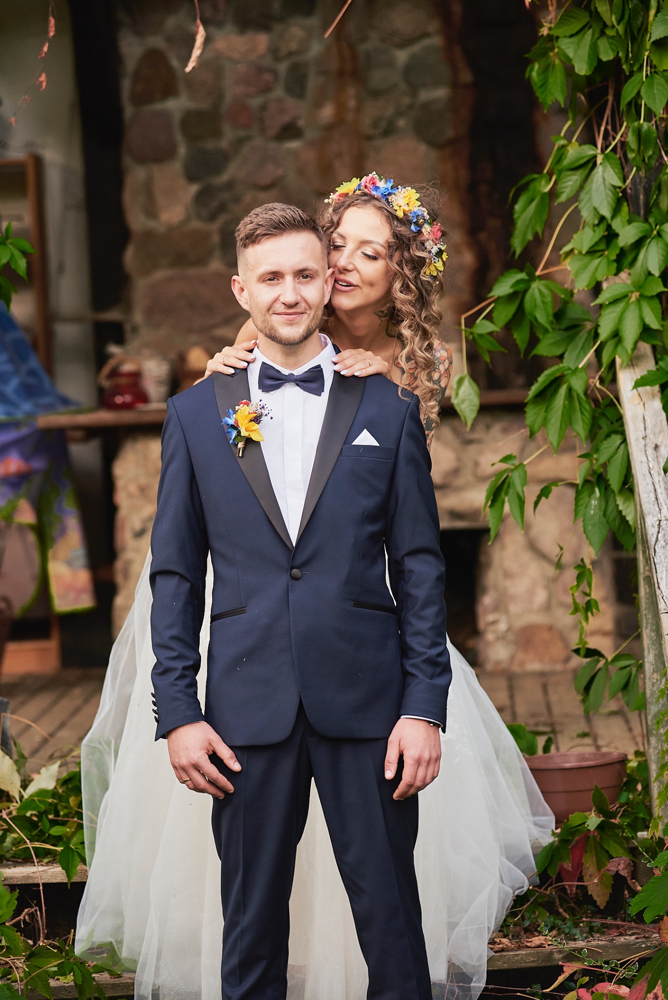 Para młoda: pan młody w granatowym smokingu z butonierką z kolorowych kwiatów, panna młoda z wiankiem na głowie obejmuje go, kamienna ściana w tle.