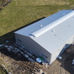 Artur Styczyrz - Zdjęcie z lotu ptaka przedstawia ukończony budynek przemysłowy z szarej blachy trapezowej z charakterystycznym świetlikiem łukowym na dachu, otoczony placem budowy z widocznymi materiałami...