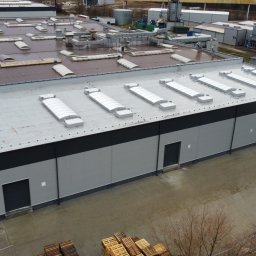 StyczyrzBuild Artur Styczyrz - Widok z góry na nowo wybudowany budynek przemysłowy z szarymi panelami, czarnym pasem i rzędem świetlików na dachu, otoczony placem z paletami i zaparkowanym samochodem.