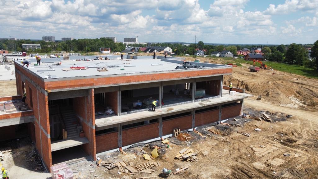 Budowa dwukondygnacyjnego budynku z cegły z widocznymi pracownikami na dachu i wewnątrz, z wykopem i sprzętem budowlanym w tle, w słoneczny dzień.