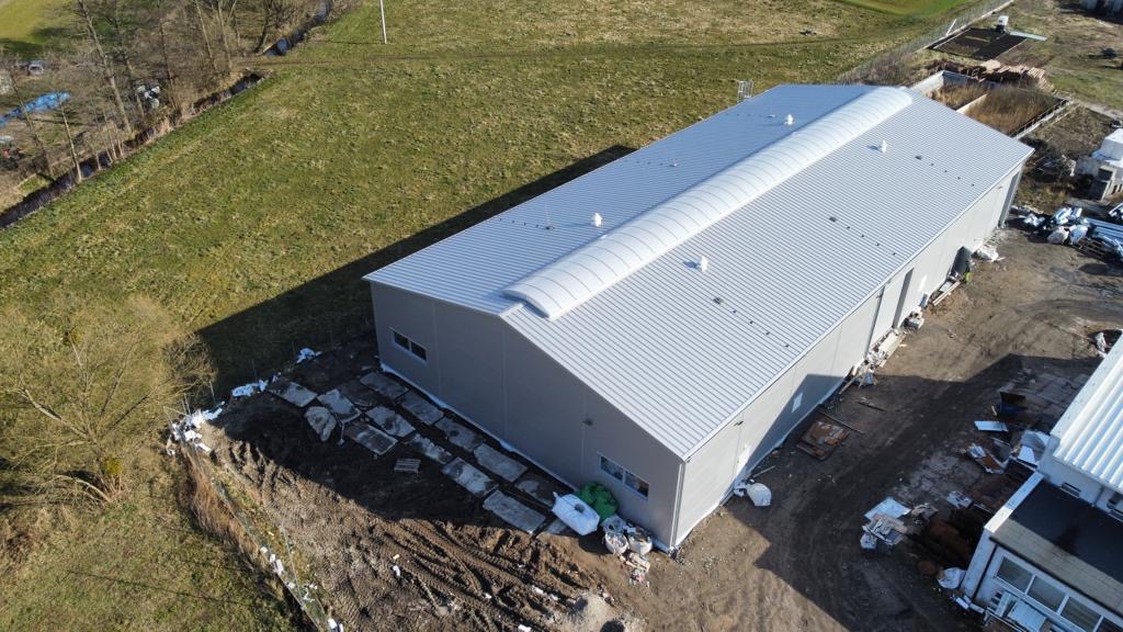 Zdjęcie z lotu ptaka przedstawia ukończony budynek przemysłowy z szarej blachy trapezowej z charakterystycznym świetlikiem łukowym na dachu, otoczony placem budowy z widocznymi materiałami...