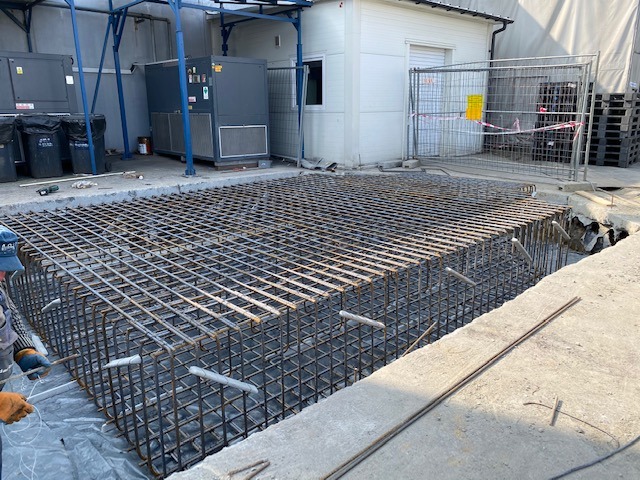 Zbrojenie fundamentu w wykopie, widoczne stalowe pręty tworzące siatkę, częściowo zalane wodą, w tle agregaty klimatyzacyjne i budynek gospodarczy.