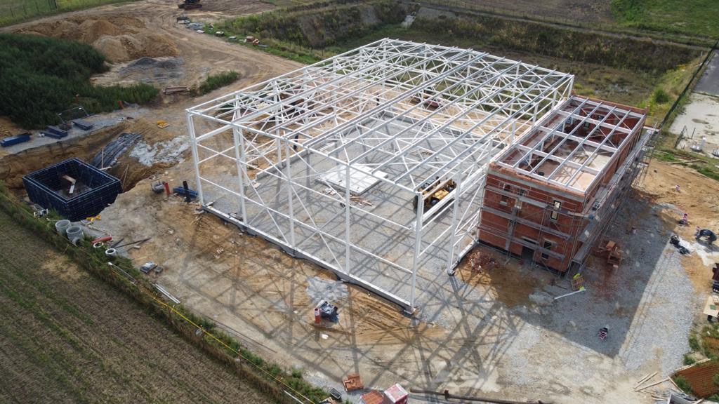 Budowa hali przemysłowej w trakcie wznoszenia konstrukcji stalowej, widoczny szkielet budynku i fragment murowanej części, wykop pod fundamenty z elementami kanalizacji, teren budowy z ziemią...