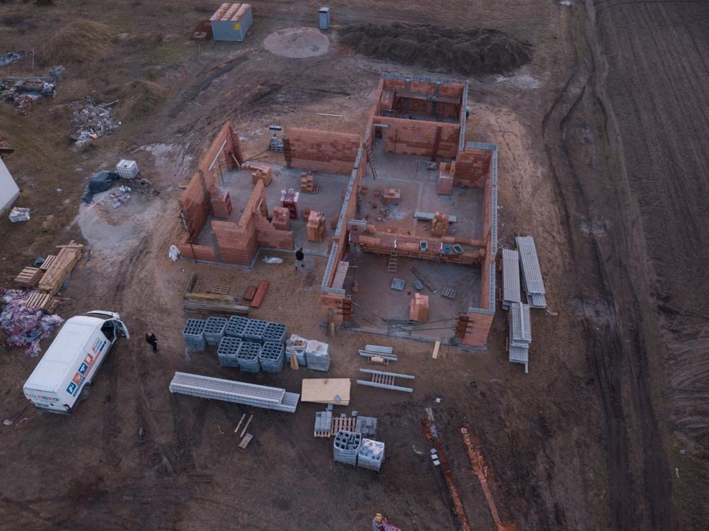 Zdjęcie z drona przedstawiające budowę domu jednorodzinnego w stanie surowym otwartym, z widocznymi ścianami z czerwonej cegły, materiałami budowlanymi na placu, oraz samochodem dostawczym firmy...