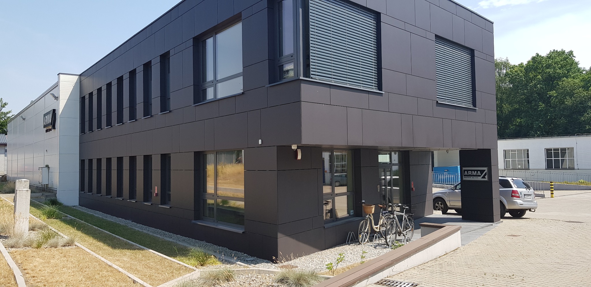 Nowoczesny budynek biurowy z ciemną elewacją panelową, widoczne rowery przed wejściem i zaparkowany samochód osobowy.