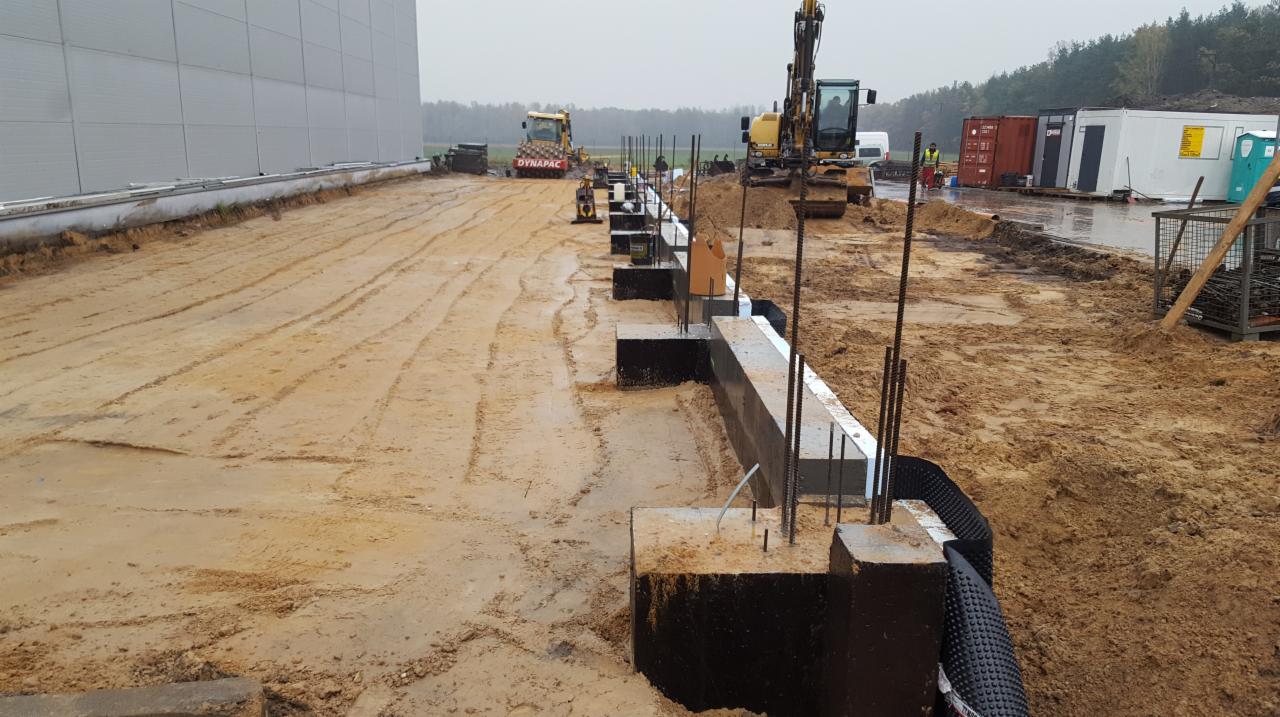 Budowa fundamentów hali przemysłowej: wylane betonowe bloki z wystającymi prętami zbrojeniowymi, widoczny ciężki sprzęt budowlany i błotniste podłoże.