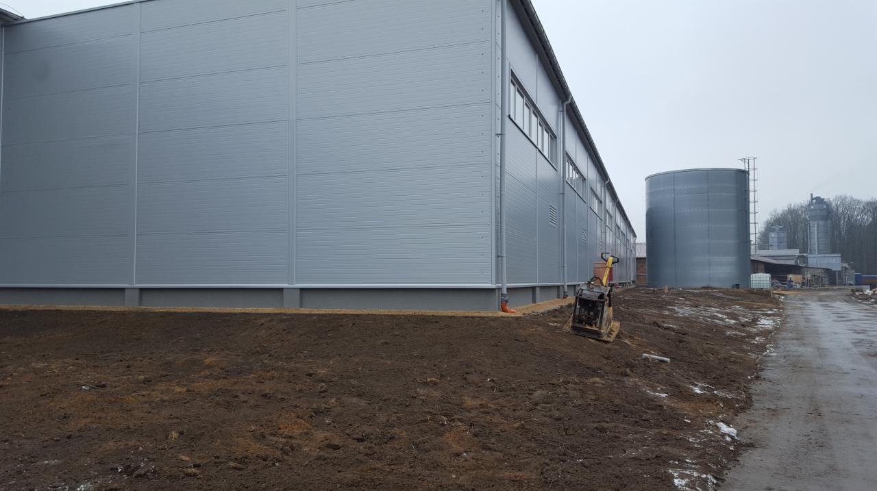 Budynek przemysłowy z szarej blachy trapezowej, widoczny zbiornik i ubijarka gruntu na nierównym terenie.