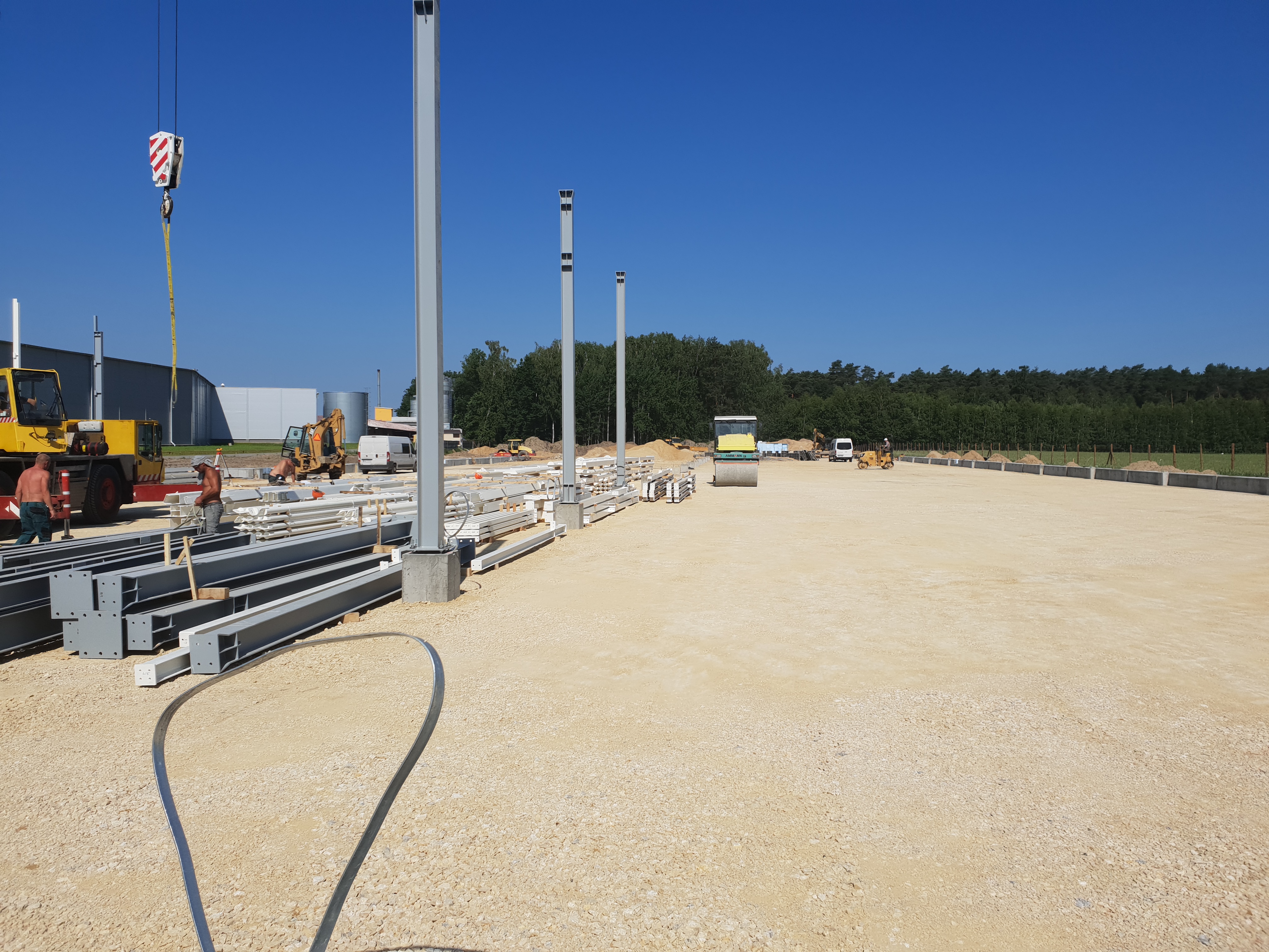 Budowa hali: widok na plac budowy z przygotowanymi stalowymi elementami konstrukcyjnymi, pracującym dźwigiem i ubijarką do gruntu na tle błękitnego nieba.