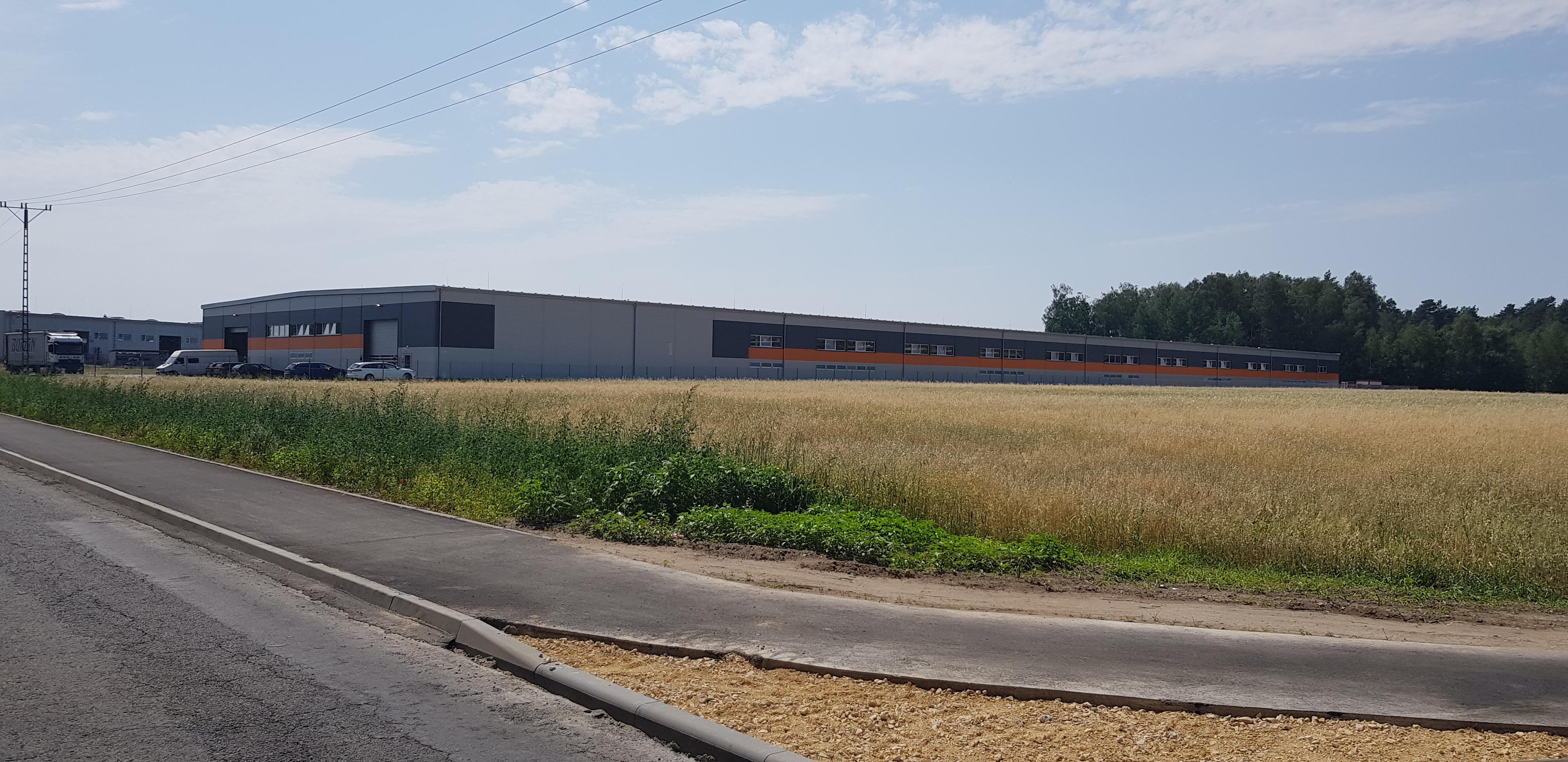 Nowoczesny budynek magazynowy z szaro-pomarańczową elewacją, widoczny z perspektywy drogi, z polem zboża na pierwszym planie.