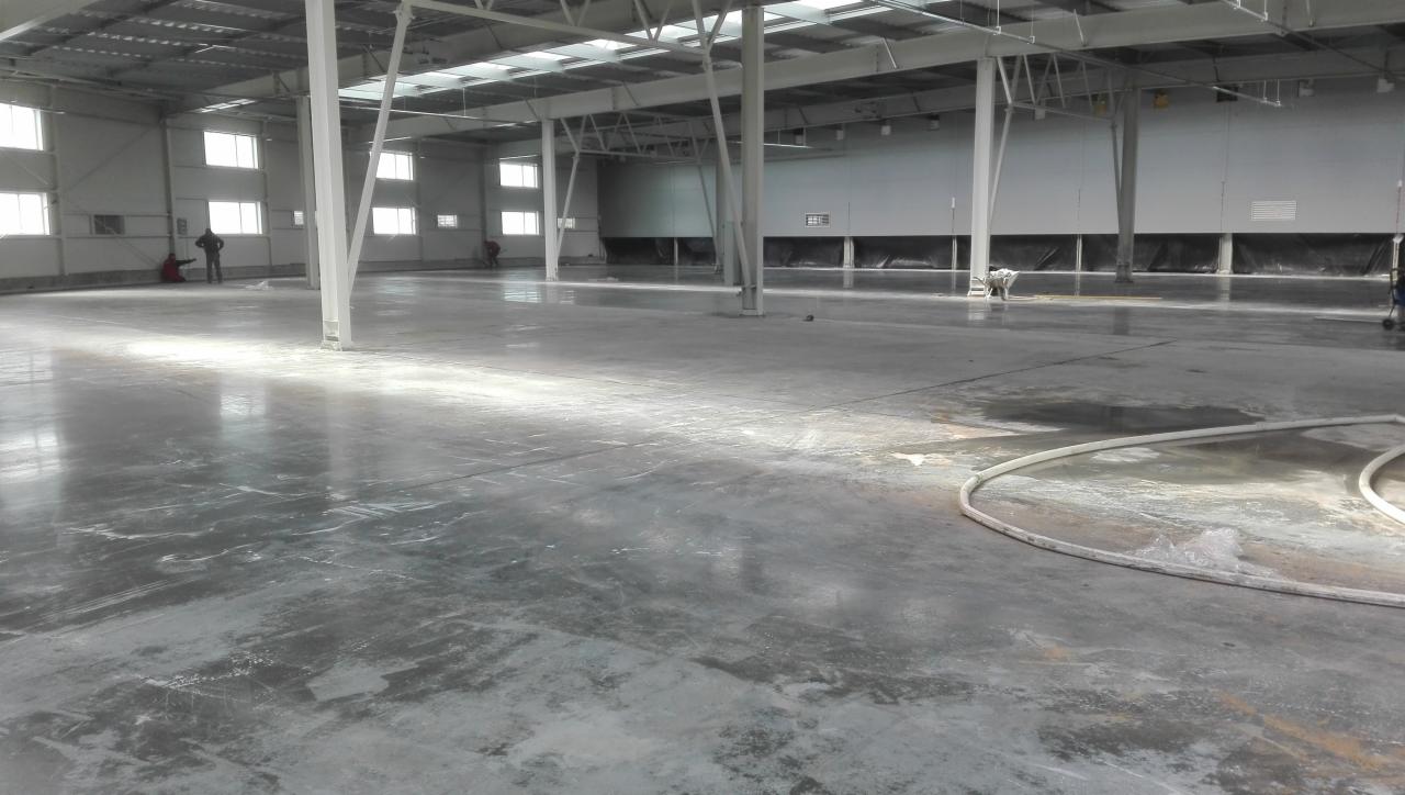 Wnętrze hali produkcyjnej z betonową posadzką w trakcie prac wykończeniowych, widoczne białe słupy konstrukcyjne, okna i osoby w oddali.