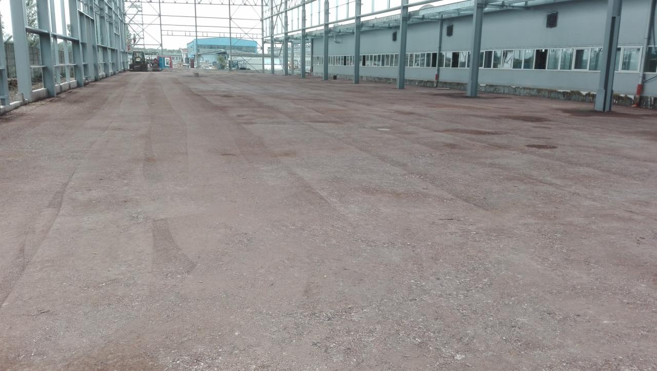 Wnętrze hali produkcyjnej w trakcie budowy, widoczne stalowe elementy konstrukcyjne, surowe podłoże i budynek w tle z rzędami okien.