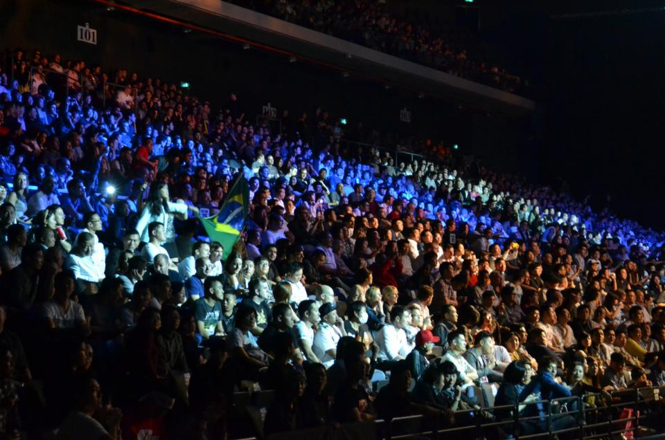 Szeroki kąt widoku na publiczność zgromadzoną w hali widowiskowej, oświetloną zróżnicowanym światłem, z widoczną flagą Brazylii.