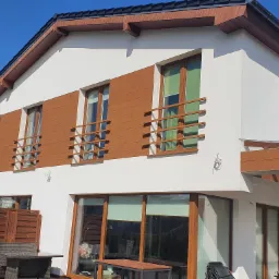 Nowoczesny dom jednorodzinny z drewnianą elewacją i prostymi, poziomymi barierkami balkonowymi w kolorze drewna. Widoczne okna z roletami, drewniane wykończenie dachu i elementy dekoracyjne...