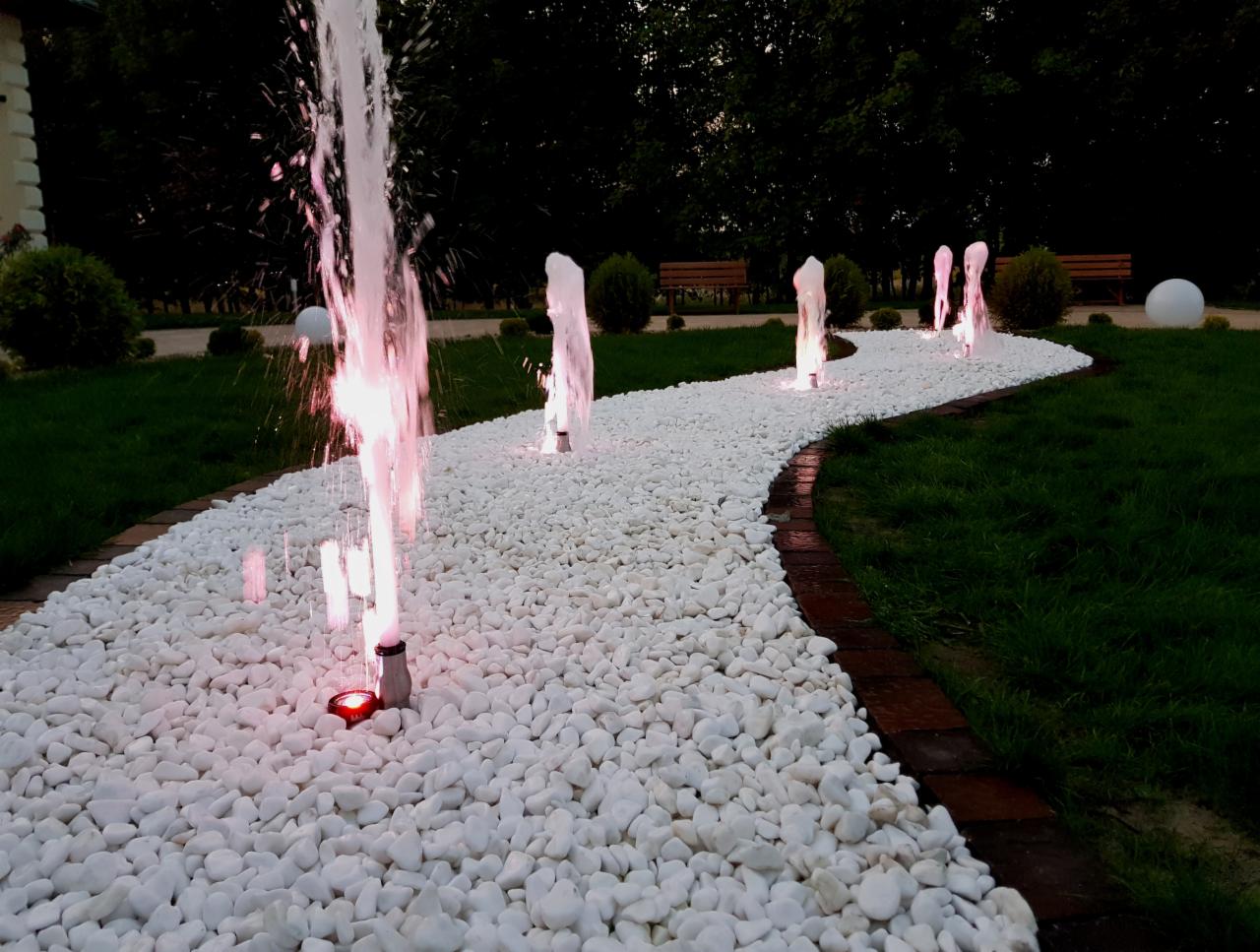 Ścieżka z białych kamieni z podświetlanymi fontannami tworzącymi szpaler w ogrodzie, oświetlona wieczorową porą.