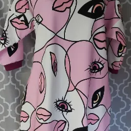 Damska sukienka z geometrycznym wzorem twarzy w odcieniach różu, bieli i czerni, wykończona bordowymi ściągaczami, prezentowana na czarnym manekinie na tle szarej tapety.