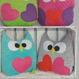 Ręcznie szyte, kolorowe poduszki dekoracyjne w kształcie sów z aplikacjami serc, umieszczone w drewnianych, bielonych skrzynkach.