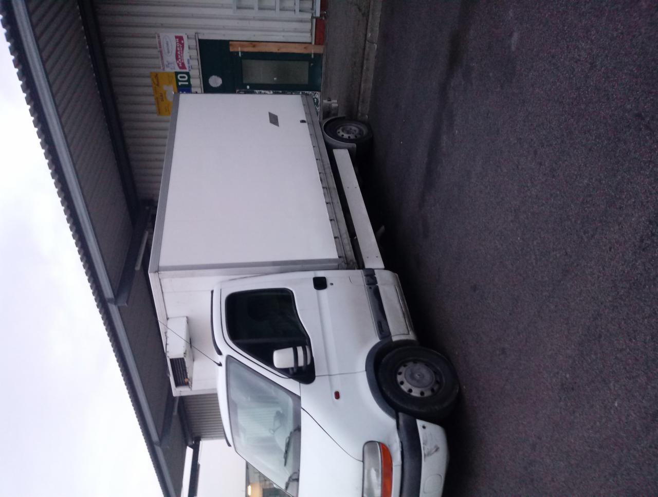 Biały samochód dostawczy typu furgon, zaparkowany na asfalcie obok budynku z szarą elewacją i zielonymi drzwiami. Widoczny agregat chłodniczy zamontowany na przedniej ścianie nadwozia.