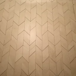 Ściana wyłożona beżowymi płytkami w jodełkę, widoczny element do poziomowania płytek w prawym górnym rogu.