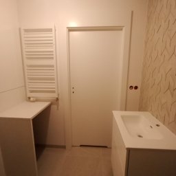 Perfectly Home - Nowoczesna, jasna łazienka z umywalką na szafce, grzejnikiem drabinkowym i ścianą z geometryczną tapetą w odcieniach beżu i brązu. Dwa gniazdka elektryczne widoczne obok drzwi.