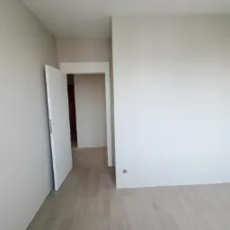 Pomalowane na jasny odcień wnętrze pokoju z otwartymi białymi drzwiami i panelami podłogowymi w odcieniach beżu i szarości.