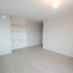 Perfectly Home - Wnętrze pokoju w trakcie prac wykończeniowych: położona podłoga z paneli, ściany częściowo pomalowane na szaro, widoczne przewody elektryczne i wiaderko z farbą przy ścianie.