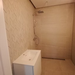 Perfectly Home - Nowoczesna łazienka z beżowymi płytkami o geometrycznym wzorze na ścianie, biała szafka pod umywalką i chromowana bateria prysznicowa z deszczownicą.