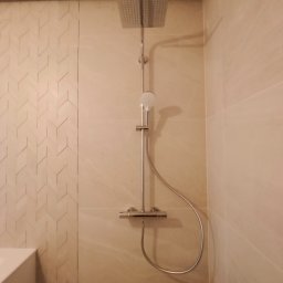 Perfectly Home - Nowoczesna kabina prysznicowa z deszczownicą, wykończona beżowymi płytkami, z akcentem ściany z mozaiką w jodełkę. Widoczny fragment białej wanny po lewej stronie.