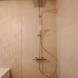 Nowoczesna kabina prysznicowa z deszczownicą, wykończona beżowymi płytkami, z akcentem ściany z mozaiką w jodełkę. Widoczny fragment białej wanny po lewej stronie.