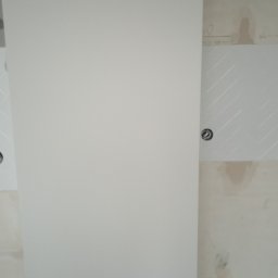 Perfectly Home - Ściana w trakcie wykańczania, częściowo pokryta białymi płytkami ułożonymi we wzór jodełki, widoczne otwory na gniazdka elektryczne, ślady po pracach budowlanych na ścianie.