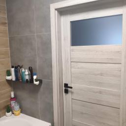 Perfectly Home - Szare płytki na ścianie łazienki, półka z kosmetykami i dekoracjami, biała umywalka oraz białe drzwi z czarną klamką.