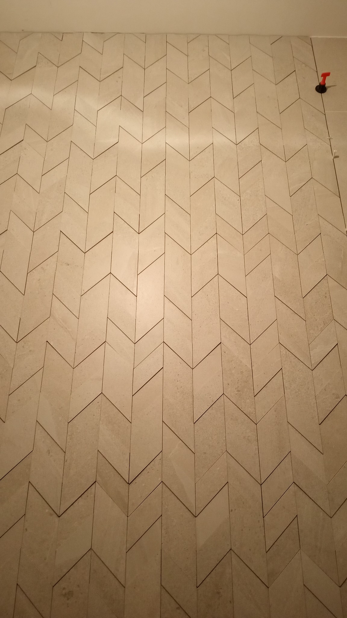 Ściana wyłożona beżowymi płytkami w jodełkę, widoczny element do poziomowania płytek w prawym górnym rogu.