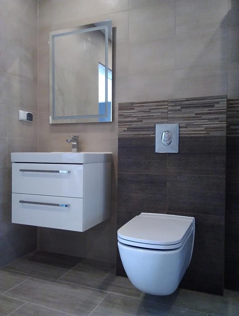 Nowoczesna łazienka z wiszącą toaletą, szafką pod umywalką i podświetlanym lustrem, wykończona płytkami w odcieniach szarości i brązu z dekoracyjnym pasem mozaiki.