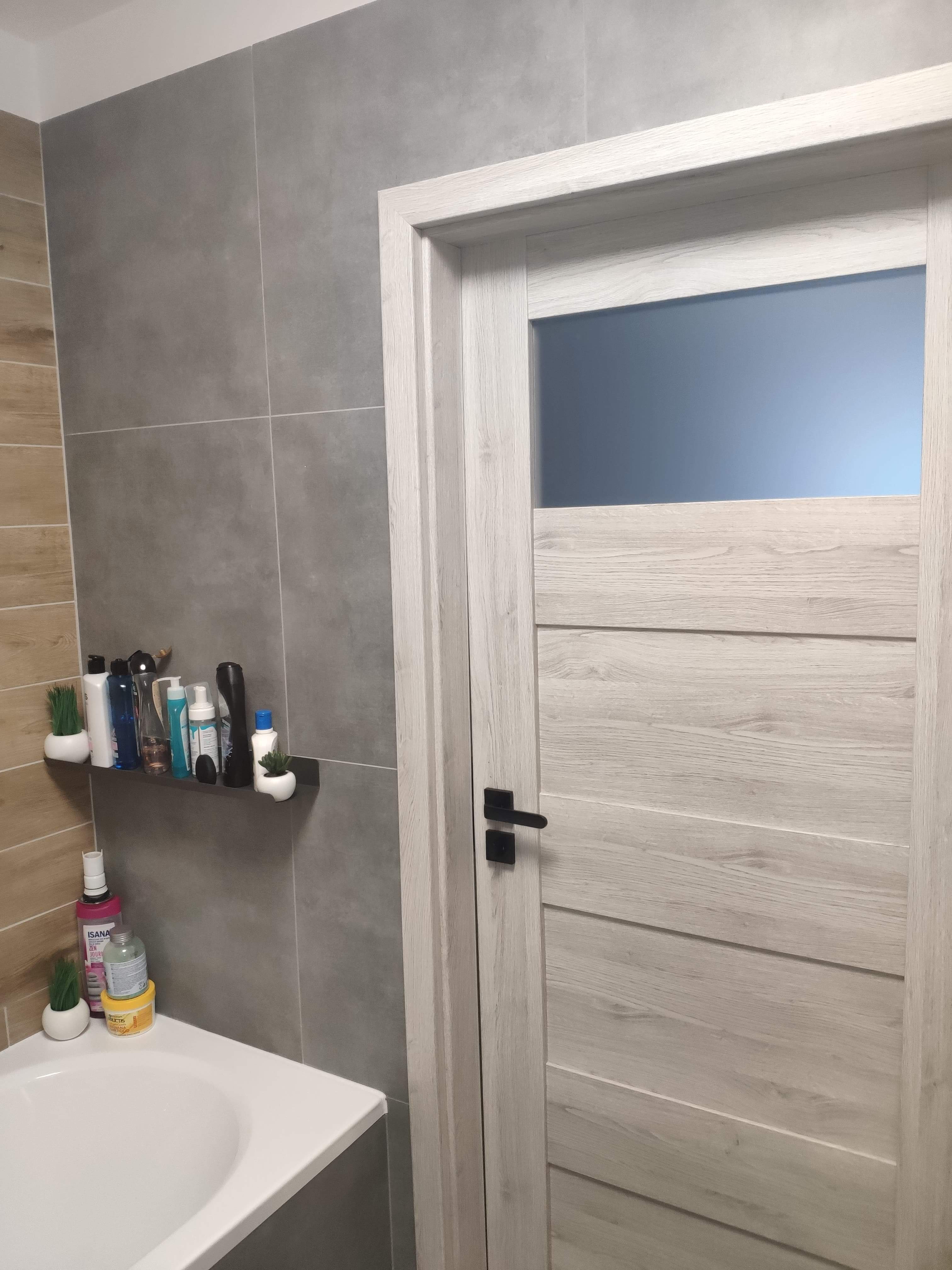 Szare płytki na ścianie łazienki, półka z kosmetykami i dekoracjami, biała umywalka oraz białe drzwi z czarną klamką.